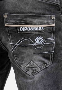 Jeans in denim nero con tasca posteriore ricamata con il marchio "CIPO&BAXX" e cuciture bianche a contrasto in un motivo decorativo.