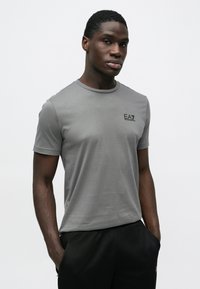 Pelēka īsroku T-krekls no gludas auduma ar apaļu kakla izgriezumu; uz krūtīm neliels melns EA7 logotips.