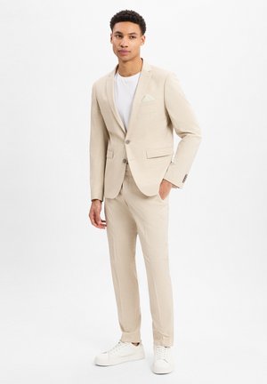 Beige suit with two buttons, notch lapels, side pockets, and a white pocket square worn over a white t-shirt and paired with white sneakers.