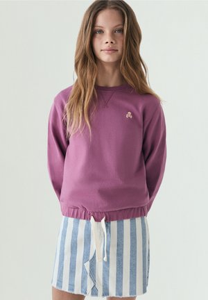 Niña joven con el pelo largo que lleva una sudadera morada con logo de calavera y una falda de rayas verticales azules y blancas con cordón ajustable.