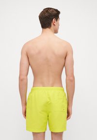 Shorts de bain jaune fluo avec ceinture élastique, une poche au dos et tissu lisse et léger. Modèle vu de dos.