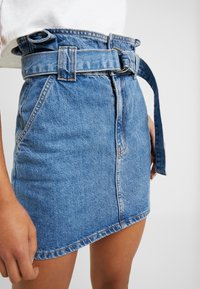 Vaalean sininen farkku mini-hame, jossa on vyöllinen vyötärö, teksturoitu kangas, ommellut yksityiskohdat ja hieman rispaantunut helma.