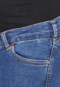 Jeans de ganga azuis com ferragens na cor cobre, costura dourada, um bolso frontal e uma textura ligeiramente usada. A cinta é visível.