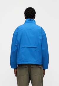 Μπλε, ελαφρύ μπουφάν με ψηλό γιακά, σχεδιασμό με χαμηλωμένους ώμους και πίσω τσέπη. Το ύφασμα φαίνεται λείο με ματ φινίρισμα.