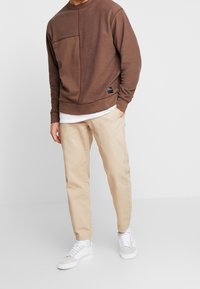 Uomo che indossa una felpa a patchwork marrone, una maglietta bianca sotto, pantaloni beige e sneakers bianche, in piedi contro uno sfondo bianco.