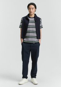 Marineblauer Strickweste über einem gestreiften Hemd in Grün, Weiß und Marineblau; dunkle Hose; weiße Sneakers; minimalistisches Design.