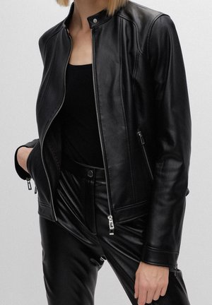 Persona que lleva una chaqueta de cuero negra sobre una camiseta negra y pantalones de cuero negros, con una mano en el bolsillo de la chaqueta y la otra relajada.