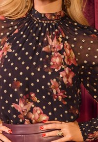 Blommig blus i genomskinlig svart tyg, med guldprickar, hög hals och smockade ärmar. Utsmyckad med rosa och lila blommönster.