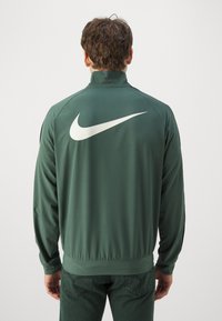 Mand iført en mørkegrøn Nike jakke med hvid swoosh-logo på ryggen, som står foran en ensfarvet lys baggrund.