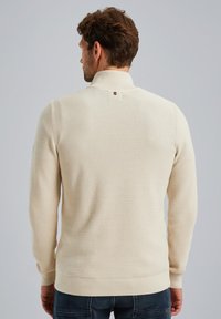 Crèmekleurige trui met een textuur, gebreid patroon, hoge kraag en een kleine logo-accent aan de achterkant. Lange mouwen en geribbelde boorden.