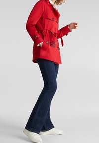 Impresionante abrigo de trench rojo con cintura ceñida, botones de doble botonadura y dos bolsillos laterales, combinado con unos vaqueros acampanados de color azul oscuro y zapatillas blancas.