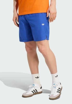 Osoba nosząca niebieskie szorty Adidas, białe skarpetki Adidas do połowy łydki, białe buty sportowe Adidas z czarnymi paskami oraz pomarańczową koszulkę.