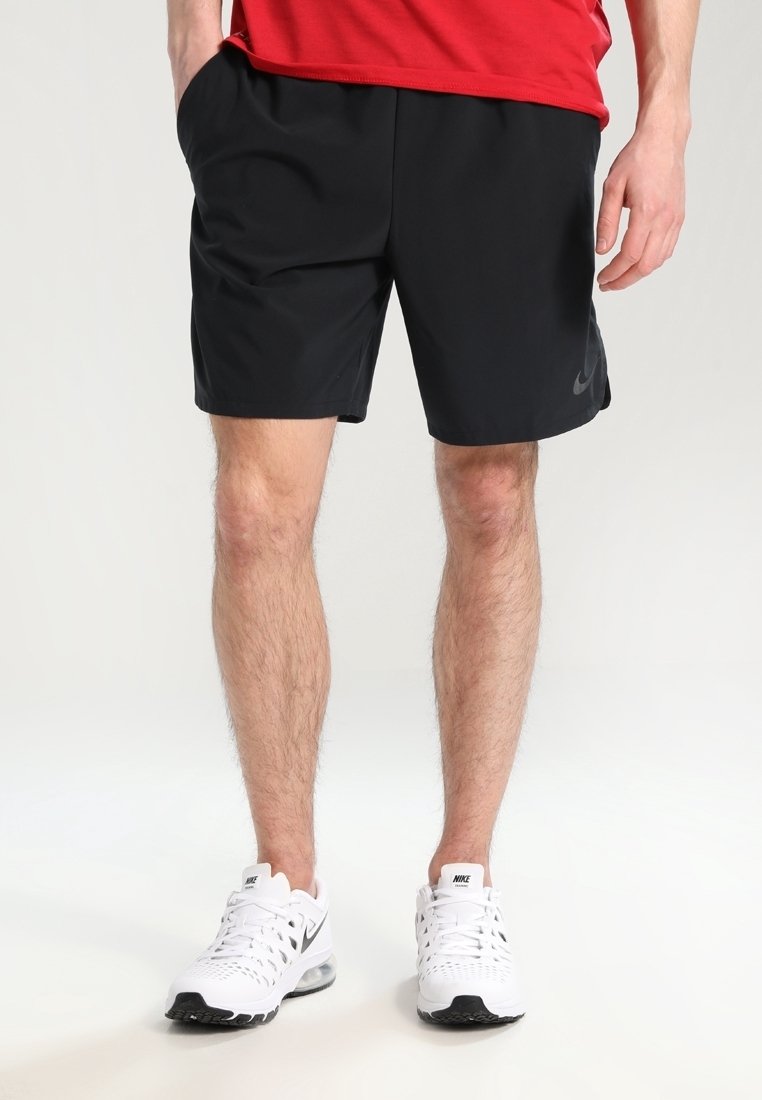 Homme portant un short Nike noir, des baskets blanches Nike et un haut rouge, debout avec une main dans la poche devant un fond uni.