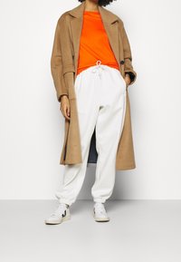 Beige Woll-Trenchcoat, orange eng anliegendes T-Shirt, weiße Jogginghose mit elastischen Bündchen und weiße Sneakers mit schwarzen Akzenten. Einfaches Design, lässige Passform.