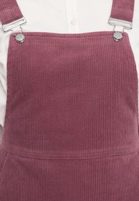 Gros plan d'une personne portant une salopette en velours côtelé rose avec des attaches à boucle métallique sur une chemise blanche boutonnée.