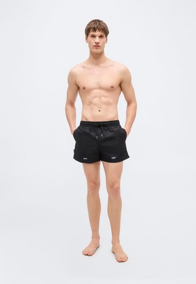 Jeune homme debout, pieds nus, portant un short noir à cordon avec le texte « OFF », mains dans les poches, sur un fond clair uni.