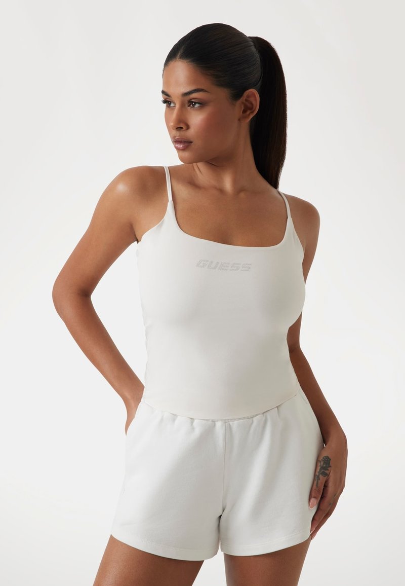 White cotton tank top with thin straps, featuring a subtly embossed "GUESS" logo. Paired with white shorts, smooth fabric texture.