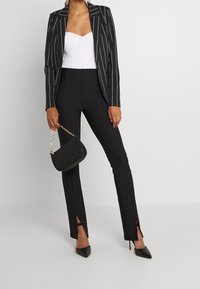 Svart kavaj med smala ränder över en vit ribbad bodysuit, kombinerad med svarta utsvängda byxor med sidorevor. Håller en liten svart handväska.