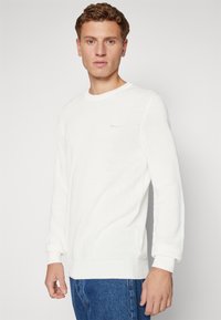Witte gebreide trui met lange mouwen, geribde manchetten en onderrand, met een subtiel logo op de borst en een gestructureerd ontwerp.