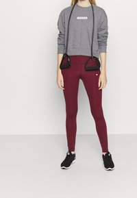 Sweatshirt gris avec logo, associé à des leggings burgundy ajustés. Des baskets de sport noires complètent la tenue sportive. Bandes ajustables attachées.