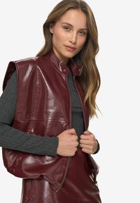 Burgundia faux nahast vest, millel on nupuvääristuse ja kõrge krae ning varrukateta disain. Kantud ribakujulise halli pika varrukaga topi peal.