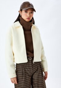 Krämmig, zippered jacka med hög krage, bärs över en brun polokrage. Texturerade bruna rutiga byxor och en brun läderkeps.
