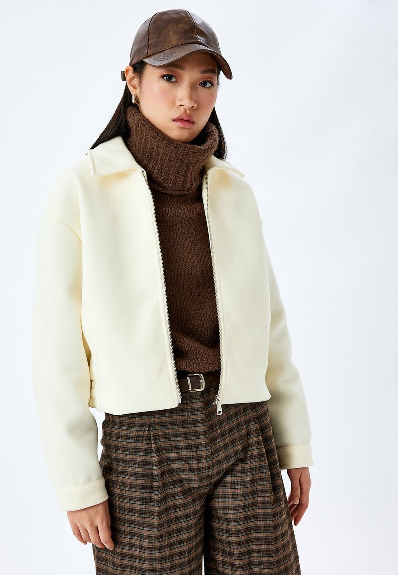 Krämmig, zippered jacka med hög krage, bärs över en brun polokrage. Texturerade bruna rutiga byxor och en brun läderkeps.