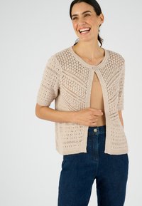 Cardigan en tricot beige clair avec des manches courtes, présentant des motifs ajourés et un col rond, associé à un jean bleu foncé.