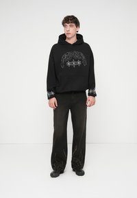 Μαύρο φούτερ με κουκούλα και μπροστινή τσέπη, με ασημένιο γραφικό σχέδιο· συνδυασμένο με σκούρο παντελόνι που διαθέτει υφασμένο μοτίβο στο τελείωμα, φορεμένο με μαύρα παπούτσια.