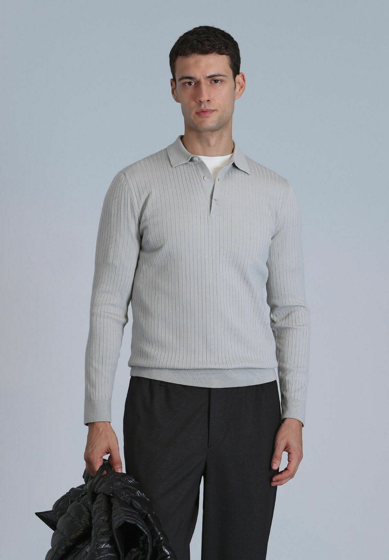 Lichtgrijze ribbelige longsleeve polo met een kraag, gedragen met donkere broeken. Het model houdt een zwarte gewatteerde jas vast.