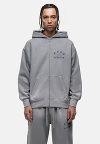 Felpa grigia con zip e tasche frontali, con la scritta in blu scuro "Good for Nothing." Tessuto morbido con vestibilità comoda e cordini regolabili.