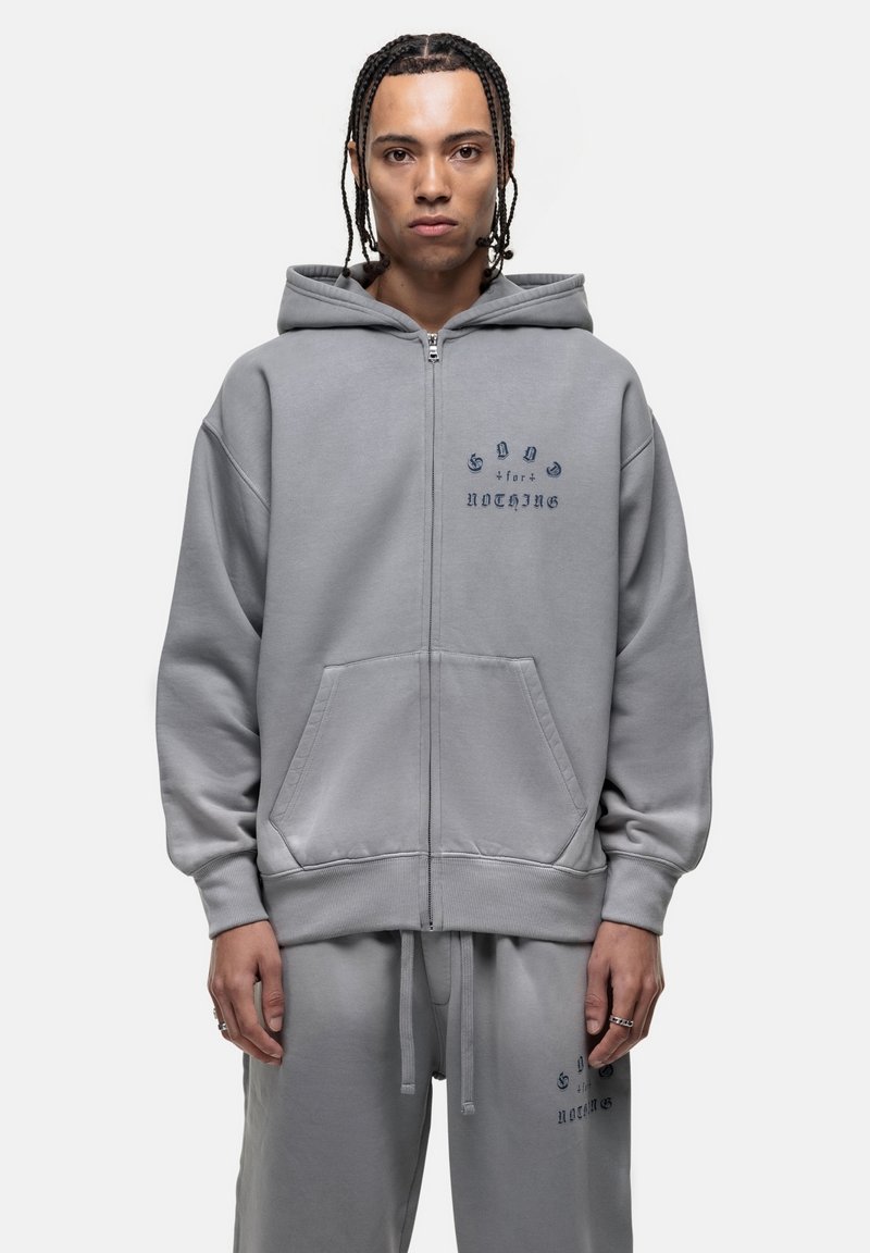 Felpa grigia con zip e tasche frontali, con la scritta in blu scuro "Good for Nothing." Tessuto morbido con vestibilità comoda e cordini regolabili.