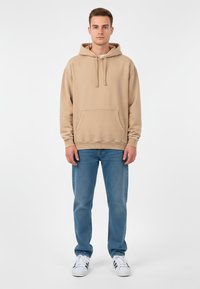 Ung man står vänd mot kameran i en beige hoodie, blå jeans och vita sneakers med svarta ränder mot en enfärgad vit bakgrund.