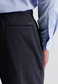 Donker getextureerde broek met een klassieke pasvorm, voorzien van een achterzak en een klein zwart label. Gecombineerd met een lichtblauw overhemd.