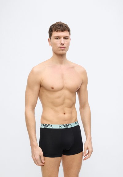 Άνδρας χωρίς πουκάμισο με κοντά σγουρά μαλλιά που φοράει μαύρα μποξεράκια με ανοιχτό μπλε ζώνη στη μέση και επώνυμα λογότυπα, στέκεται μπροστά σε ένα απλό φόντο.