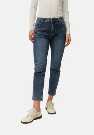 Mujer con vaqueros azules de cintura alta y corte cropped, camiseta blanca de manga larga y zapatillas color crema con rayas rosas, parada frente a un fondo liso.