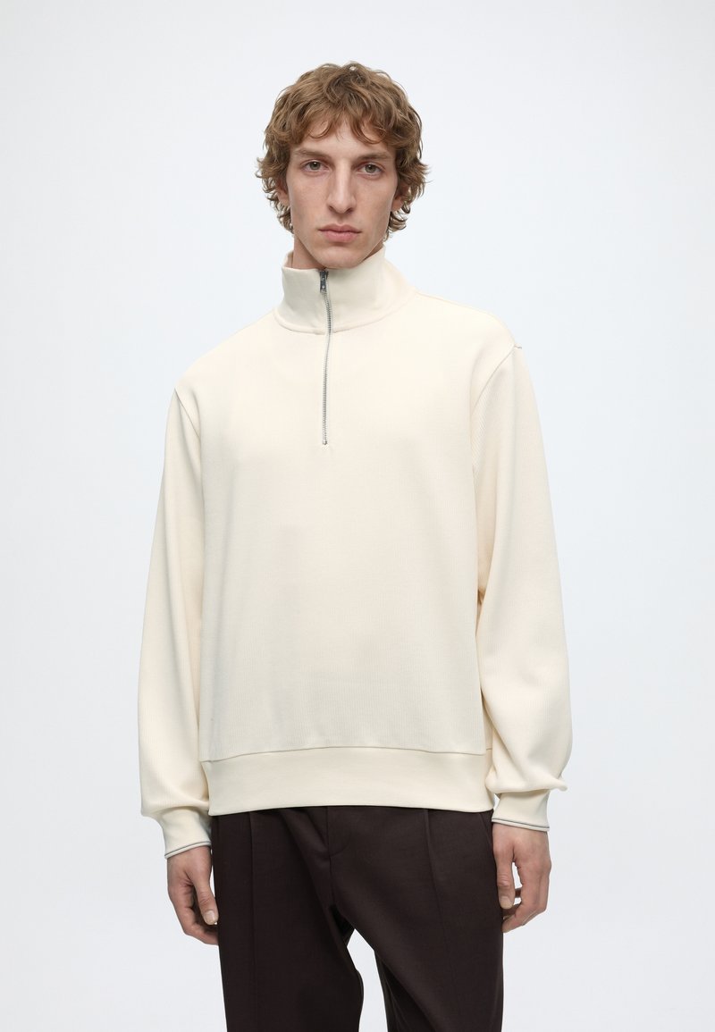 Jeune homme aux cheveux bouclés porte un pull crème à col montant zippé et un pantalon foncé, debout devant un fond clair uni.