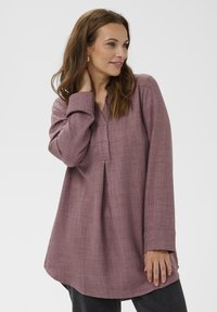 Ženska s polovico dolžine rjavih las, oblečena v ohlapno mauve tuniko in temne hlače, pozira s roko na vratu proti enotni ozadju.