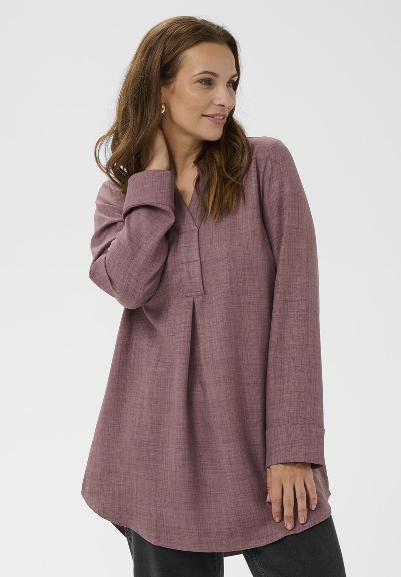 Ženska s polovico dolžine rjavih las, oblečena v ohlapno mauve tuniko in temne hlače, pozira s roko na vratu proti enotni ozadju.