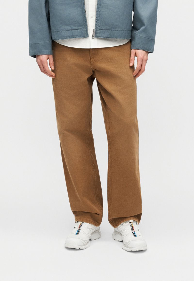 Personne portant un pantalon marron ample, des baskets blanches avec des détails noirs et bleus, une chemise blanche et une veste zippée bleue.