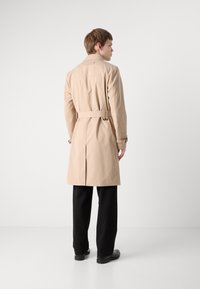 Trench coat bej cu talie cu curea, fanta din spate singulară și bretele cu nasturi pe mâneci, asortat cu pantaloni negri și pantofi.