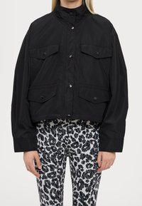 Veste noire raccourcie avec col haut, fermeture à boutons sur le devant et deux grandes poches poitrine. Portée sur des leggings noirs et blancs à motifs.