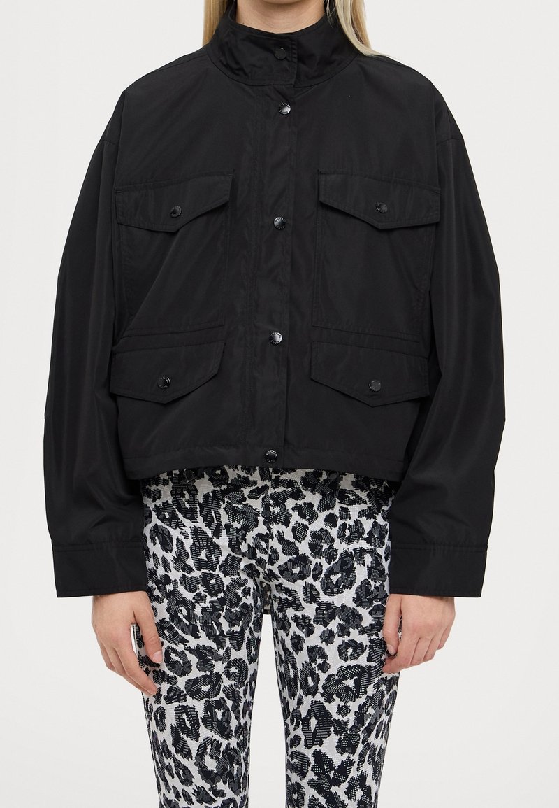 Veste noire raccourcie avec col haut, fermeture à boutons sur le devant et deux grandes poches poitrine. Portée sur des leggings noirs et blancs à motifs.