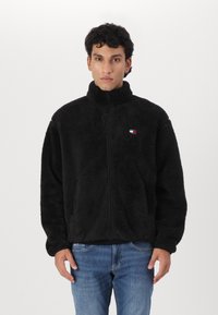 Chaqueta de forro polar negra con cuello alto y cierre de cremallera. Presenta un pequeño logo bordado en el lado izquierdo del pecho. Tejido suave y texturizado, corte relajado.