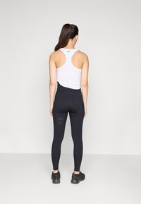 Hvit racerback singlet, svart høyt liv leggings med teksturerte detaljer, og svarte treningssko, sett fra baksiden, mot en ensfarget bakgrunn.
