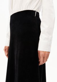 Schwarzer Samt-Midi-Rock mit glatter Textur, tailliertem Bund und einem seitlichen Schlitz. Kombiniert mit einem weißen Hemd mit langen Ärmeln und Knöpfen.