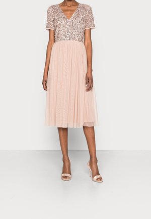 Kvinde iført en knælængde, lyserød kjole med en paillet-bodice med V-udskæring og en plisseret tyl-nederdel, kombineret med matchende åbne hæle.