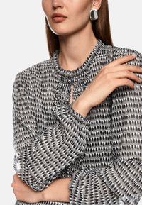 Graues gemustertes Blusenshirt mit Rüsche am Hals und an den Ärmelbündchen, das ein gepunktetes Design in Schwarz und Weiß aufweist. Die Hand des Models ruht elegant auf dem Arm.