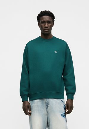 Mand iført en grønblå crewneck-sweatshirt med lille hvid Adidas-logo og lyseblå jeans med blå grafiske detaljer på højre ben.