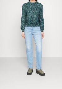 Blommig långärmad topp i gröna och svarta nyanser, tillsammans med ljusblå jeans och mörkgröna slip-on skor med tjocka sulor.